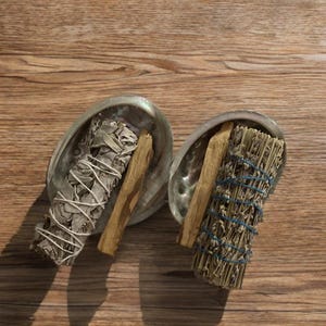 May include: Two abalone shells each hold a bundle of sage and a piece of palo santo wood. One sage bundle is wrapped in white string, the other in blue string. The background is a wooden surface.