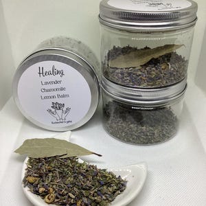 May include: Three glass jars and a small white dish filled with dried herbs and flowers. The jars have silver lids and labels that read "Healing," with the ingredients listed as lavender, chamomile, and lemon balm. A bay leaf rests on top of the herbs.