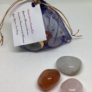 May include: A navy blue mesh bag with gold stars and a drawstring, containing crystals. A tag reads "Cancer 6/21-7-22 Crystal Kit" and lists crystal properties. Three polished stones are in front: orange, gray, and pink.