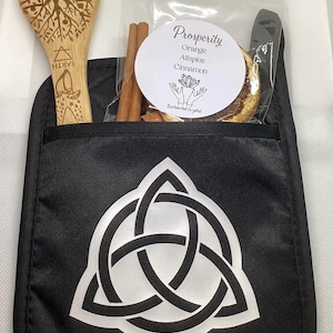 May include: A black pot holder with a white Celtic knot design, holding a wooden spoon with a tree and moon carving, a bag with cinnamon sticks, and a label that reads "Prosperity" with ingredients.