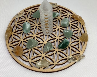 May include: A wooden, circular decorative piece with a geometric pattern, surrounded by various green and amber-colored gemstones. A clear quartz crystal with moon phase engravings stands in the center.
