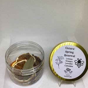 May include: A clear jar filled with dried orange slices, bay leaves, and other botanicals, next to a gold-rimmed tin with the text "Ostara/ Spring Renewal" and a bee illustration. The tin lists ingredients: Lemons, Oranges, Lavender, and Calendula.
