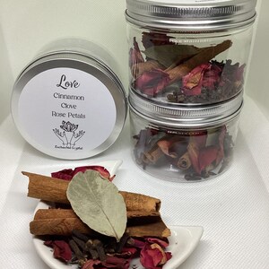 May include: Three clear jars and a tin with silver lids, filled with cinnamon sticks, rose petals, cloves, and a bay leaf. The tin and jars have labels with the word "Love" and ingredient names. A small white dish holds similar ingredients.