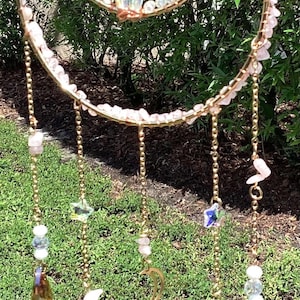 May include: A celestial-themed suncatcher with a gold-colored metal frame. The design features a crescent moon, stars, and crystals in shades of pink, clear, and iridescent colors. The suncatcher is suspended by a gold chain.
