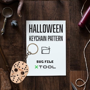 May include: A white card with the text "HALLOWEEN KEYCHAIN PATTERN" and "SVG FILE XTOOL" printed on it. A brown leather Halloween mask keychain with a gold metal ring is in the foreground.