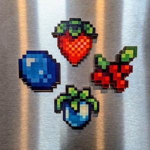 May include: Four pixel art fruit magnets made of wood. The set includes a red strawberry, a blue blueberry, a dark red raspberry cluster, and a blue berry with green leaves. Each piece features a distinct blocky, digital-inspired design.