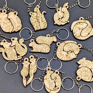 May include: A collection of light-colored wooden keychains featuring laser-engraved animal pairs, including cheetahs, rabbits, and raccoons. Each keychain consists of a wooden animal cutout attached to a silver-toned metal split ring by a chain.