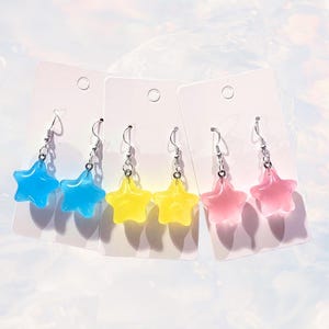 May include: Four pairs of star-shaped dangle earrings. The earrings are made of clear resin with a translucent color. The colors are blue, yellow, and pink.