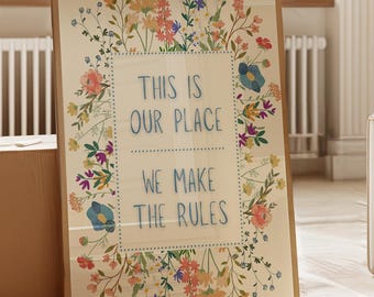 以下が含まれることがあります： 「THIS IS OUR PLACE WE MAKE THE RULES」のテキストが花柄の縁で囲まれた額入りのプリント。木製の床に、大きな植物と段ボール箱の隣に表示されています。パレットの色は、ソフトパステルと自然な木の色合いが含まれています。