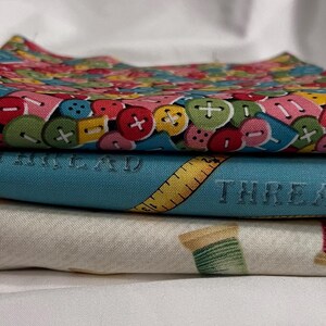May include: A stack of four fabric squares with different patterns. The top square has a colourful button pattern. The second square is blue with a yellow measuring tape and the word "THREAD" printed on it. The third square has a white background with a repeating pattern of spools of thread.