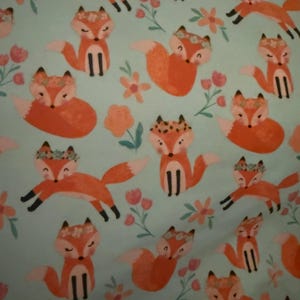 May include: A repeating pattern of orange foxes wearing floral crowns and flowers on a light blue background. The foxes are in various poses, with some standing and others running. The flowers are in shades of pink and orange.