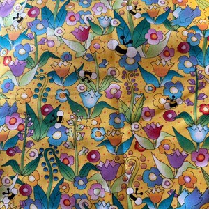 May include: A vibrant yellow fabric with a colorful floral pattern. The design features various flowers in shades of purple, blue, pink, and orange, along with green leaves and cartoon bees. The overall pattern is dense and detailed.