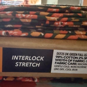 May include: A stack of fabric with a fall leaves pattern in shades of green, orange, and brown. The label reads "Interlock Stretch" and lists fabric details, including a width of 57 inches.