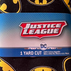May include: Black fabric featuring repeating yellow Batman logos. A blue and white label displays the Justice League logo, the DC logo, and the text "1 Yard Cut". The fabric is 36in x 44in.