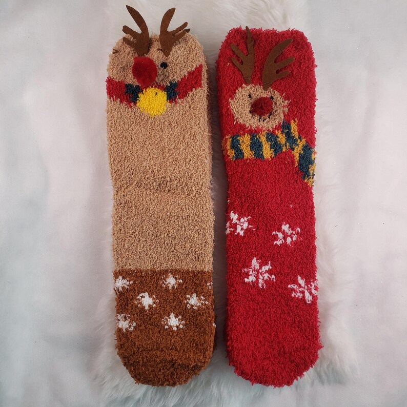 May include: Two pairs of fuzzy Christmas socks with reindeer designs. One pair is brown with a red pom-pom nose and a bell. The other pair is red with a snowflake pattern.