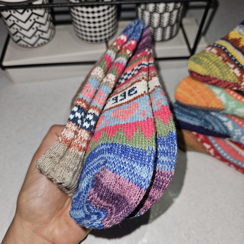 May include: A pair of colorful knitted socks with a geometric pattern. The socks are folded in half and have a beige, blue, pink, and green color scheme. The socks have the word "BEE" written on them.