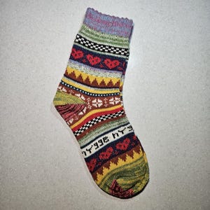 May include: A colorful striped sock with a pattern of hearts and geometric shapes. The sock is made of a soft, knitted fabric and has a light blue, green, yellow, red, brown, and black color scheme.