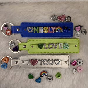Pode incluir: Três chaveiros de couro com diferentes pingentes e letras. O chaveiro superior é azul com a palavra "ONESLY" em pedrarias. O chaveiro do meio é verde com a palavra "LOVE" em pedrarias. O chaveiro inferior é prateado com as palavras "I LOVE YOU" em pedrarias.