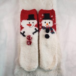 May include: Pair of white fuzzy socks with red and white snowman designs. The snowmen have black eyes, black buttons, and black hats.