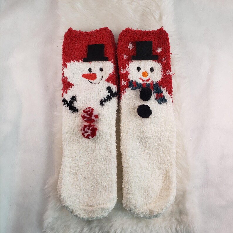 May include: Pair of white fuzzy socks with red and white snowman designs. The snowmen have black eyes, black buttons, and black hats.