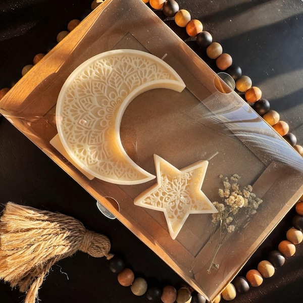 May include: A decorative candle set featuring a crescent moon and a star, both with intricate, carved patterns. The candles are a creamy off-white color and are presented in a brown box with dried flowers. A beaded strand and tassel are also visible.