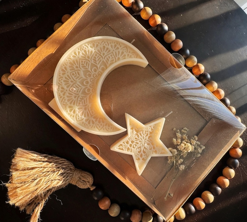 May include: A decorative candle set featuring a crescent moon and a star, both with intricate, carved patterns. The candles are a creamy off-white color and are presented in a brown box with dried flowers. A beaded strand and tassel are also visible.