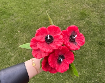 May include: A bouquet of red poppy-shaped candles with white wicks. The candles are held in a hand wearing a black leather bracelet with a gold charm.