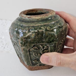 May include: A small, antique, green ceramic jar with a wide mouth and a textured surface. The jar has embossed floral patterns and a brown base. The jar is held in a hand.