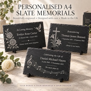 Puede incluir: Tres placas conmemorativas rectangulares de pizarra A4 con bordes naturales irregulares, cada una sobre un soporte negro. Las superficies de piedra gris oscuro presentan grabados blancos con nombres, fechas y motivos decorativos de rosas, una cruz y notas musicales.