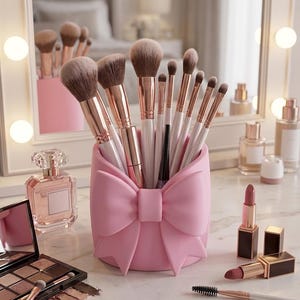 May include: A collection of makeup brushes in a pink bow-shaped holder. The brushes have light-colored bristles and rose gold handles. Other makeup items, including lipsticks, eyeshadow, and perfume, are also visible.
