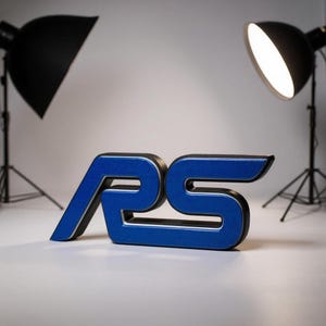 May include: A blue and black RS emblem, possibly for a car. The letters are in a stylized font with a metallic blue finish and a black outline. The emblem is on a white surface, lit by studio lights.