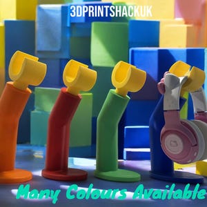 以下が含まれることがあります： レゴブロックの形をしたカラフルなヘッドホンスタンド4つと、青いスタンドにピンクのヘッドホンが置かれています。背景には「3DPRINTSHACKUK」というテキストが表示されています。前景には「Many Colours Available」というテキストが表示されています。