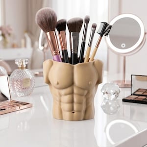 May include: A beige, sculpted torso-shaped makeup brush holder filled with various makeup brushes. The holder has defined abdominal muscles. The brushes have black, gold, and pink handles and soft bristles. A lighted vanity mirror and makeup are in the background.