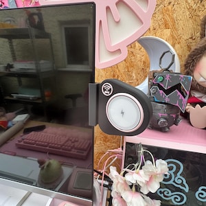 以下が含まれることがあります： 黒とピンクのワイヤレス充電器がコンピューターモニターに取り付けられています。充電器は白い充電面とピンクのトリムを備えています。背景には、ピンクのキーボード、装飾用のピンクのリボン、小さなロボットの置物が含まれています。