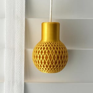 May include: A gold-colored, decorative blind pull with a lattice design. The pull is suspended by a white cord against a white background. The pull has a cylindrical top and a spherical, diamond-patterned body.