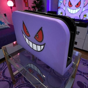 May include: A purple games console case with a cartoon Gengar face, red eyes and white grin. The case sits on a glass table with a gold frame, reflecting the Gengar design. A TV in the background shows a Gengar image.