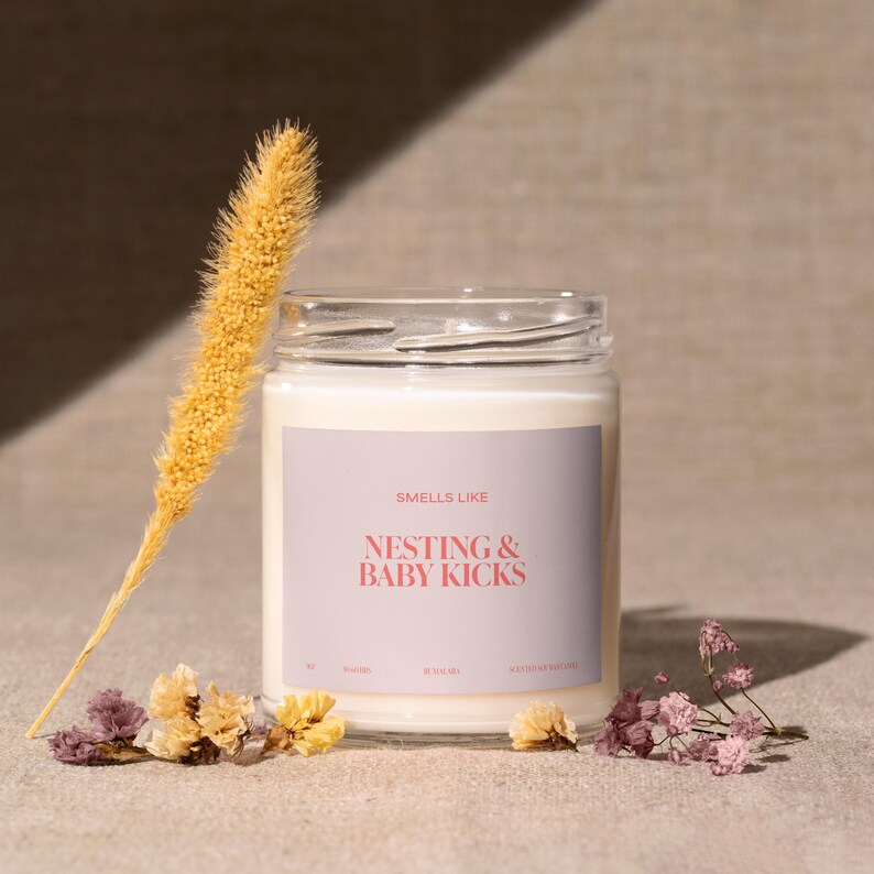 May include: A white candle in a glass jar with a grey label that reads "Smells Like Nesting & Baby Kicks". The candle is surrounded by dried flowers.