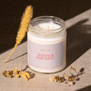 May include: A white candle in a clear glass jar with a grey label that reads "Smells Like Nesting & Baby Kicks". The candle is surrounded by dried flowers.