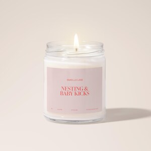 May include: A clear glass candle jar with a white candle inside. The label on the jar says "Smells Like Nesting & Baby Kicks".