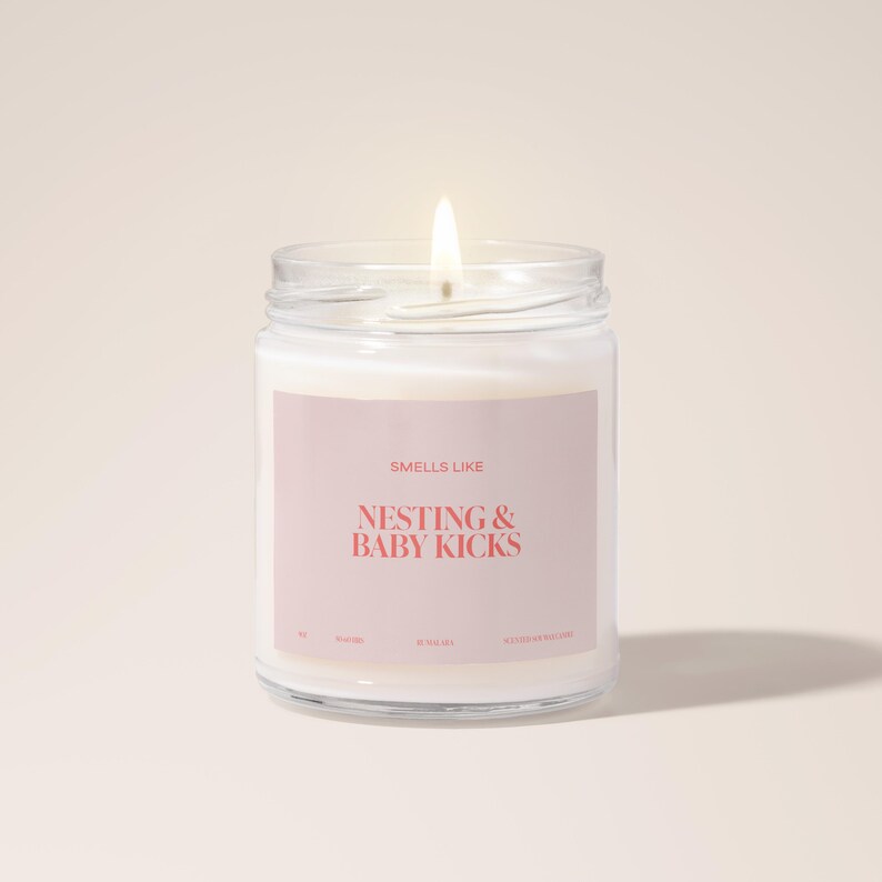 May include: A clear glass candle jar with a white candle inside. The label on the jar says "Smells Like Nesting & Baby Kicks".