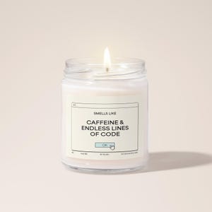 Peut inclure: Une bougie parfumée allumée dans un bocal en verre transparent. La bougie porte une étiquette blanche avec le texte "Smells Like Caffeine & Endless Lines of Code" et une icône de curseur de souris d'ordinateur. La bougie est sur fond rose clair.