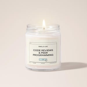 Könnte beinhalten: Eine weiße Kerze in einem Glasgefäß mit einem Etikett, auf dem "Smells Like Code Reviews & Peer Programming" mit einem "OK"-Button-Grafik steht.