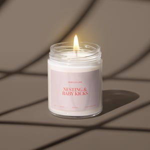 May include: A white candle in a clear glass jar with a label that reads "Smells Like Nesting & Baby Kicks". The candle is lit and the flame is visible.