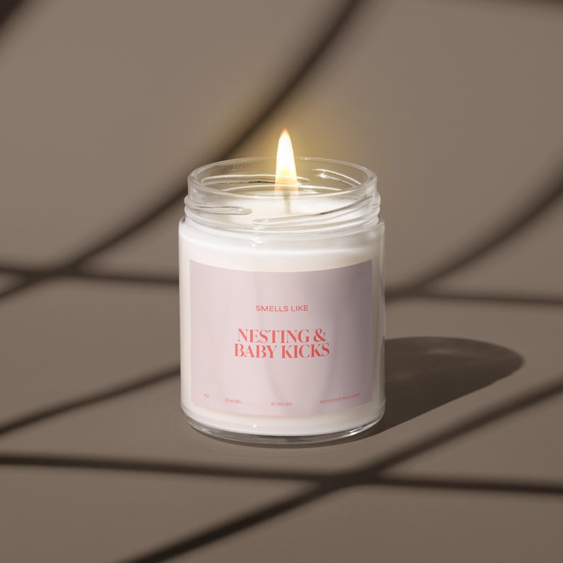 May include: A white candle in a clear glass jar with a label that reads "Smells Like Nesting & Baby Kicks". The candle is lit and the flame is visible.