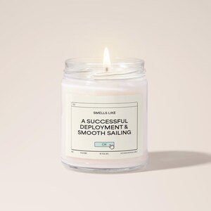 以下が含まれることがあります： 「Smells Like A Successful Deployment & Smooth Sailing」とラベルが貼られたガラスの瓶に入った白いキャンドル。キャンドルには「OK」ボタンのグラフィックが描かれている。