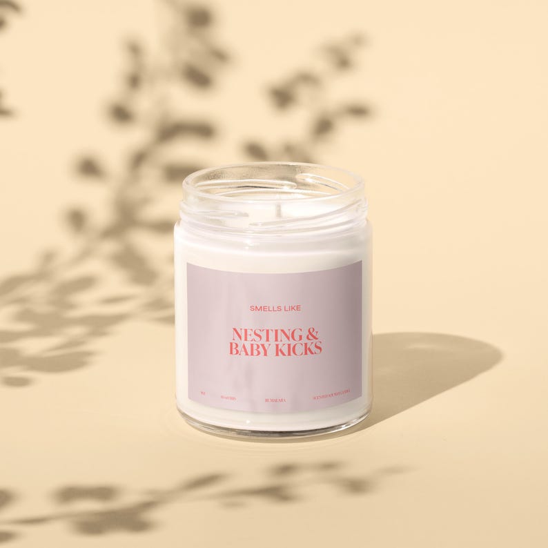 May include: A white candle in a clear glass jar with a pink and white label that says "Smells Like Nesting & Baby Kicks".