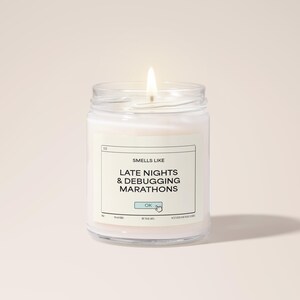 Könnte beinhalten: Eine weiße Kerze in einem Glasgefäß mit einem Etikett, auf dem "Smells Like Late Nights & Debugging Marathons" mit einem "OK"-Button-Grafik steht.