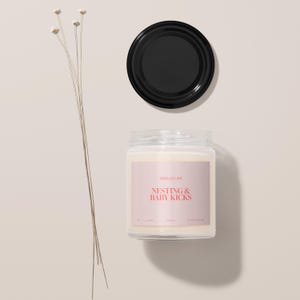 May include: A white candle jar with a pink label that says "Smells Like Nesting & Baby Kicks". The candle is on a white surface with a black lid next to it.