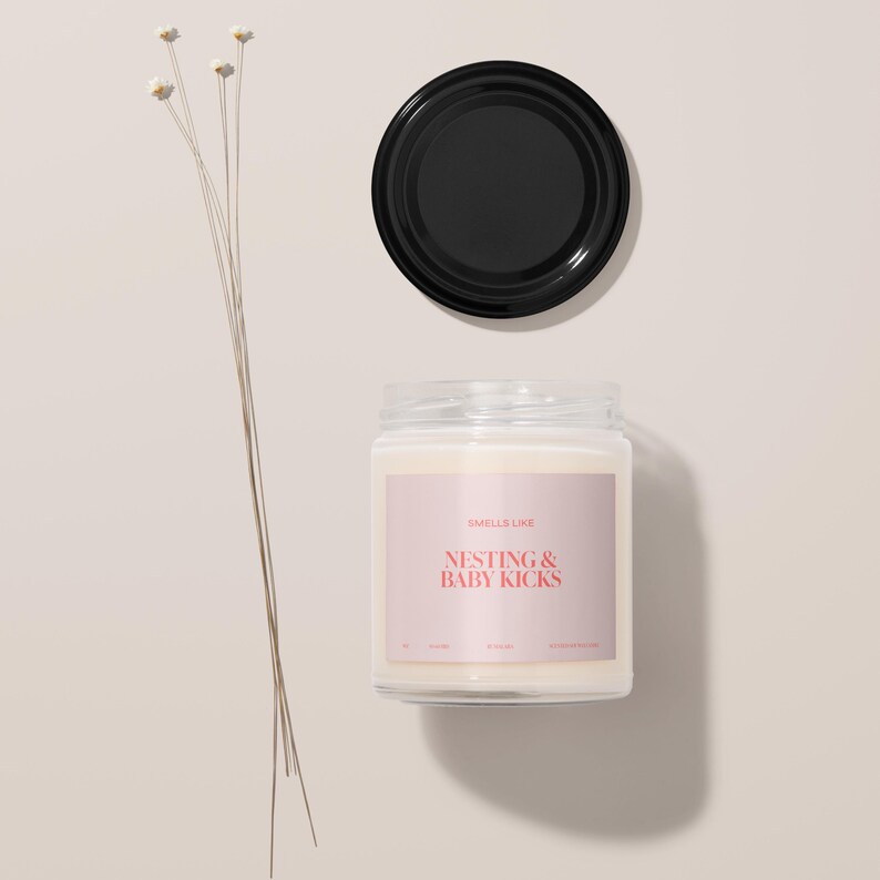 May include: A white candle jar with a pink label that says "Smells Like Nesting & Baby Kicks". The candle is on a white surface with a black lid next to it.