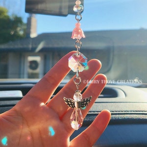 May include: A car charm featuring a clear crystal angel with silver wings, a faceted crystal, and pink flower and bead accents. The charm hangs from a silver chain and is suspended in front of a bright blue sky.